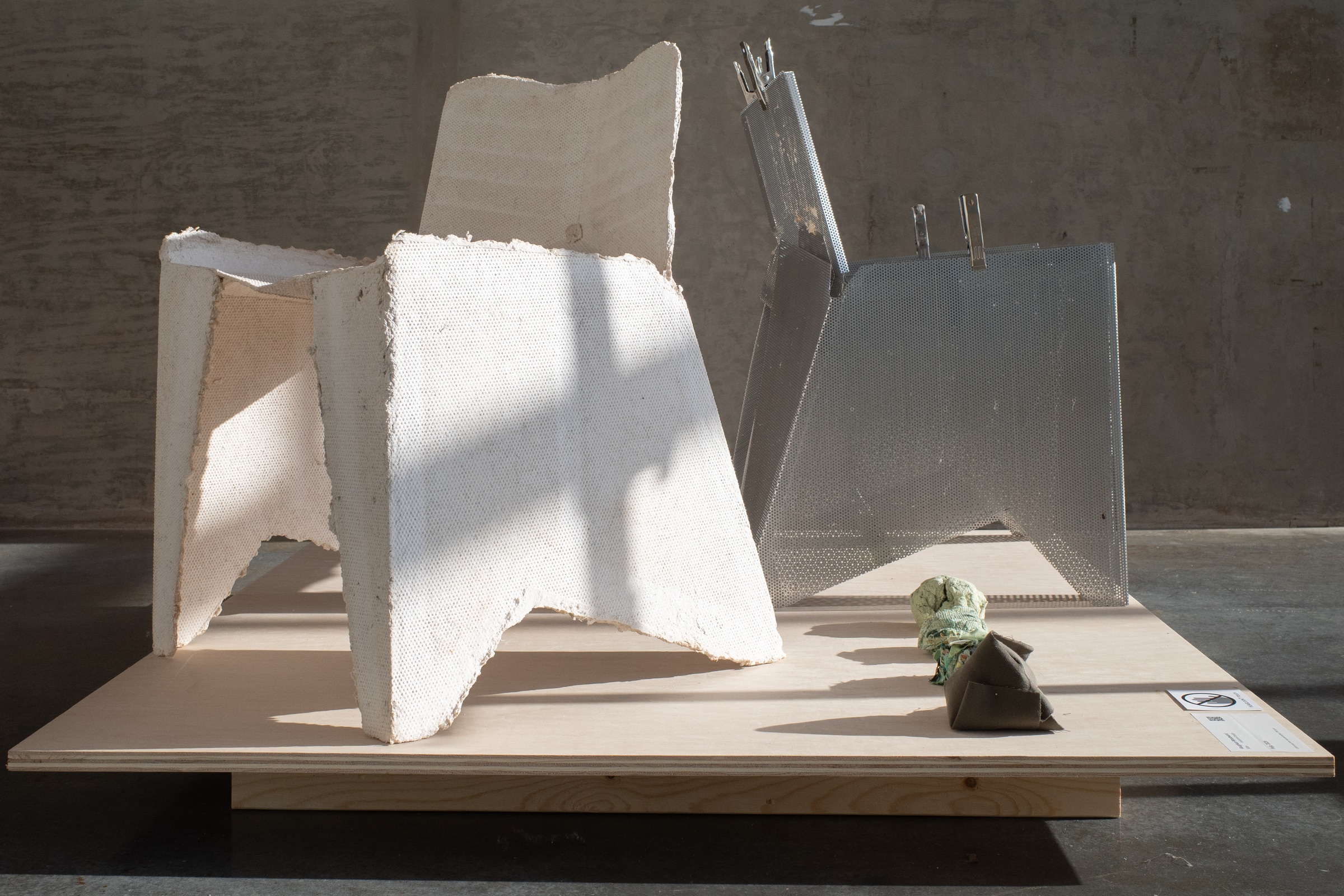 Two sculptural chairs displayed on a wooden platform—one made of rough white material resembling plaster and the other of perforated metal—lit by natural light in a gallery setting.