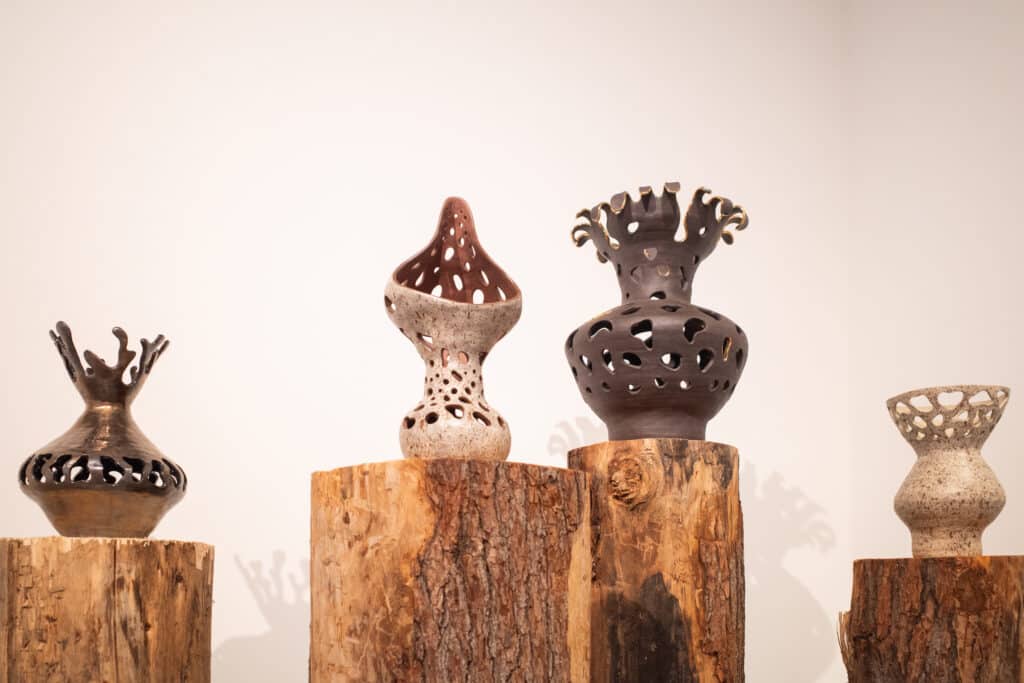 Four ceramic vessels with cut-out patterns and organic shapes displayed on tree stump pedestals against a plain wall.