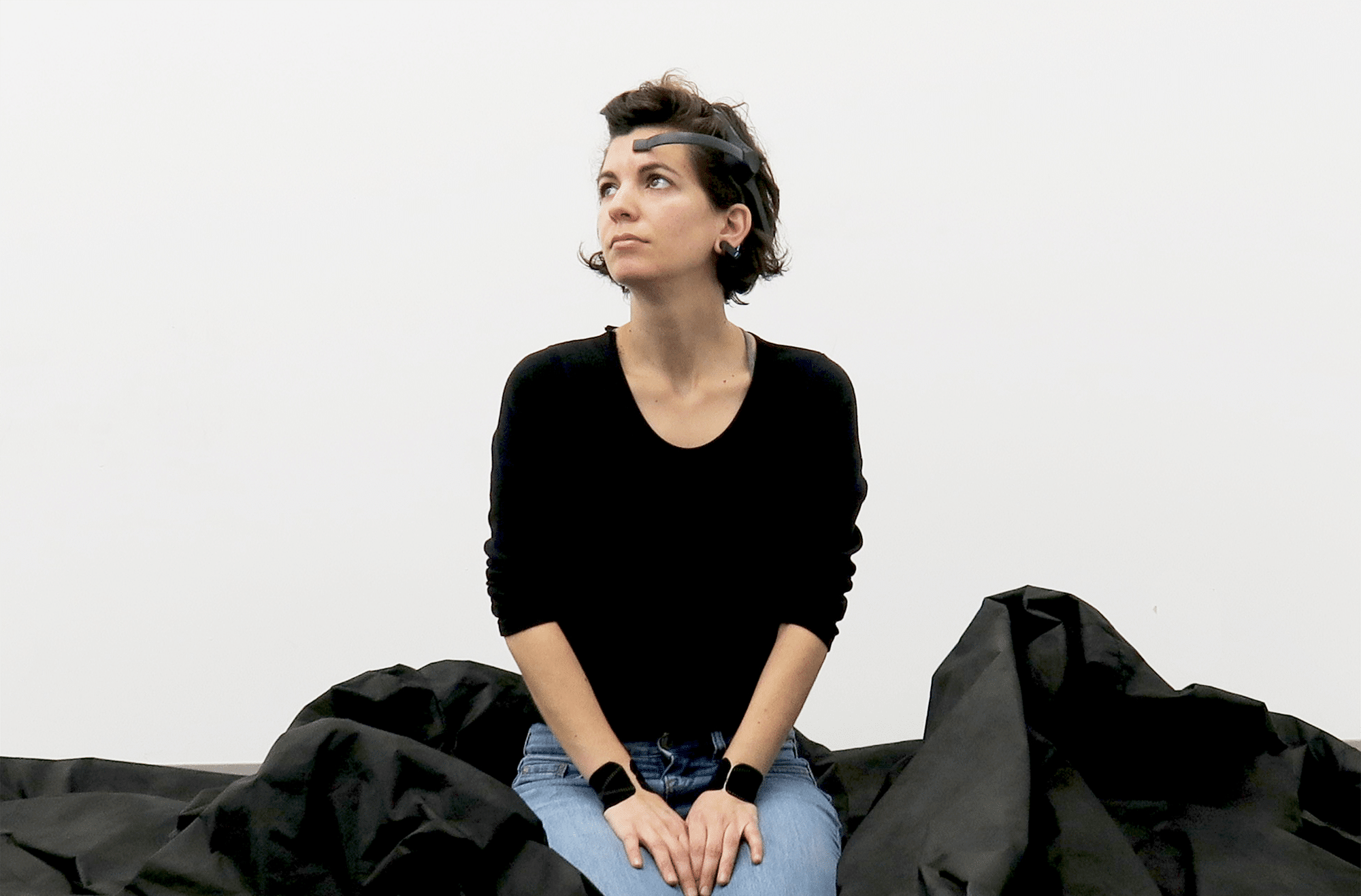 Person seated on the floor in front of a white wall, wearing a black long-sleeve shirt and blue jeans, surrounded by large folds of black fabric, looking slightly upward and to the side.