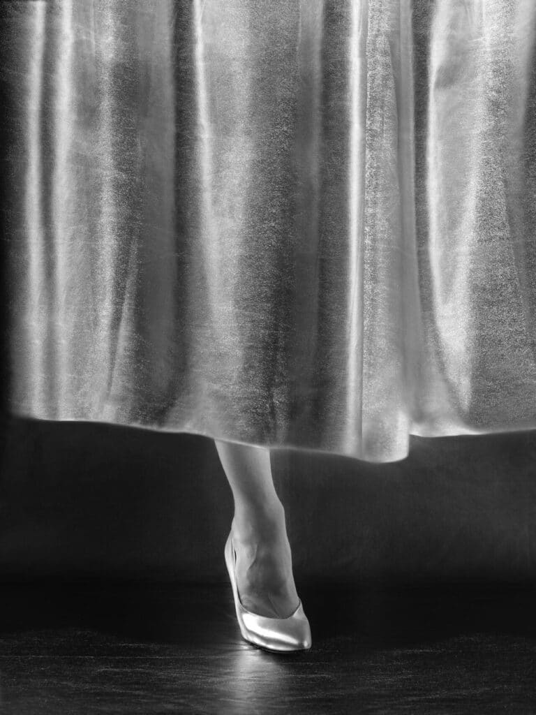 A close-up photo of a person’s legs wearing shiny heels, partly concealed by a metallic curtain or gown.