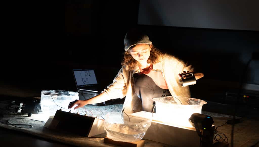 A student kneels on the floor during a performance, adjusting wires and shining a small light over large translucent bowls filled with water and ice, with a glowing effect. A laptop and audio equipment sit nearby.