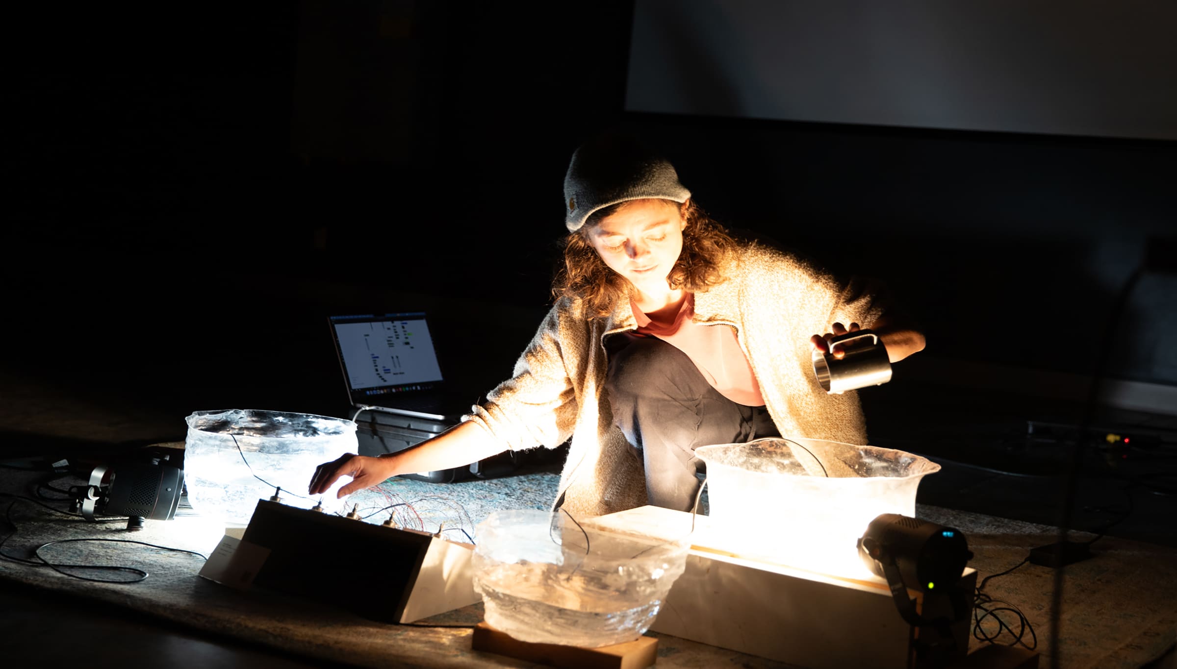 A student kneels on the floor during a performance, adjusting wires and shining a small light over large translucent bowls filled with water and ice, with a glowing effect. A laptop and audio equipment sit nearby.