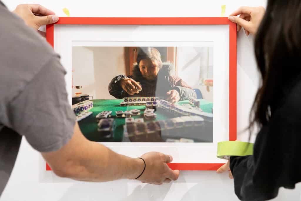 Two people hang framed photo in a bright orange frame. The photo fills most of the frame; all we see are the hands and arms of the people hanging it.