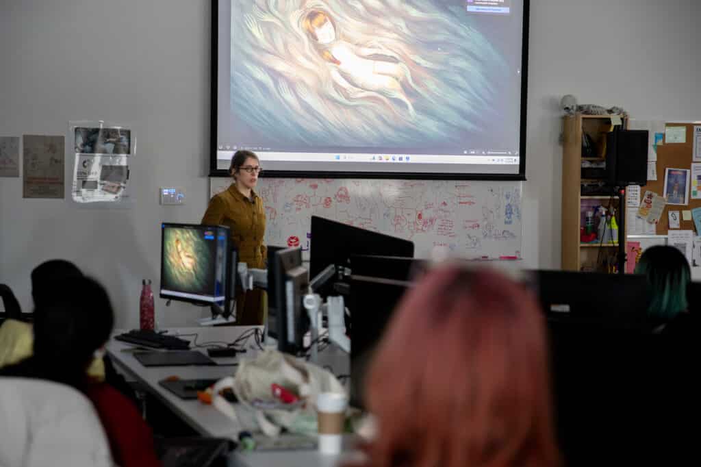An instructor stands at the front of a classroom giving a presentation on digital art. A large screen displays an illustration of a child lying in swirling, colorful patterns. Students sit at computer workstations in the foreground, and a whiteboard with doodles and notes is visible behind the instructor.