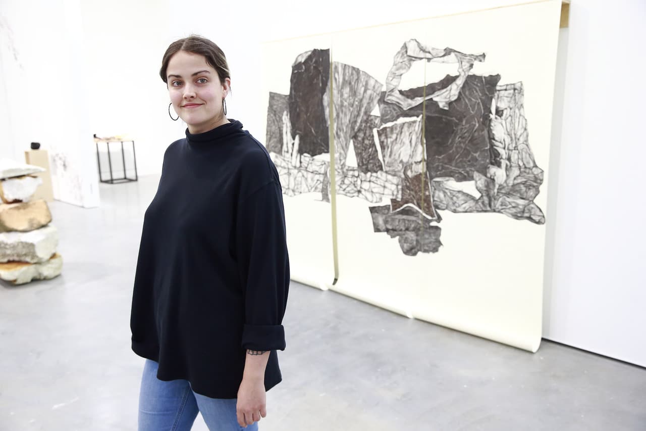 An artist stands in a gallery beside a large black-and-white drawing, with sculptures and other works displayed in the background.