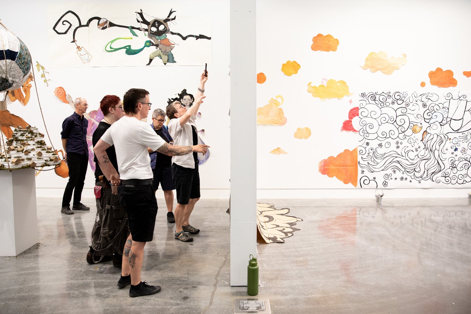 We are looking at an exhibition space, bisected by a wall. One side has several people looking at something on their side of the wall. The other side is empty. The walls facing us have colourful artwork and sculpture on and around them.