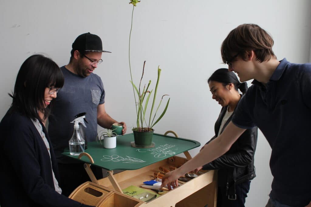 Four people gathered around a wooden cart with plants and gardening tools, smiling and interacting with the setup.