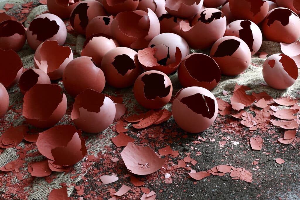 Broken red egg shells scattered across a burlap cloth and dark surface, with fragments and pieces spread around.
