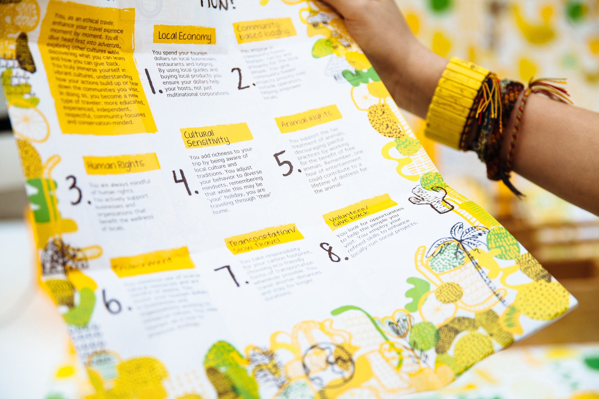 Close-up of a person holding a printed sheet with illustrated, colourful text and graphics about travel ethics.