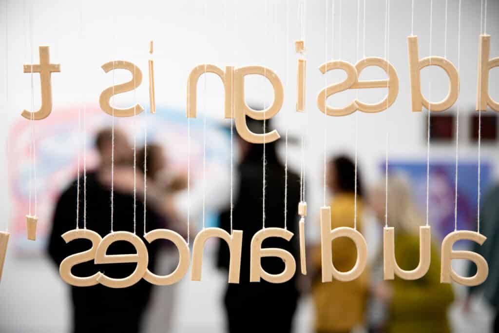 Close-up of suspended wooden letters hanging from strings, spelling words backward as they are viewed from behind. People are blurred in the background, engaged in conversation in a gallery space.