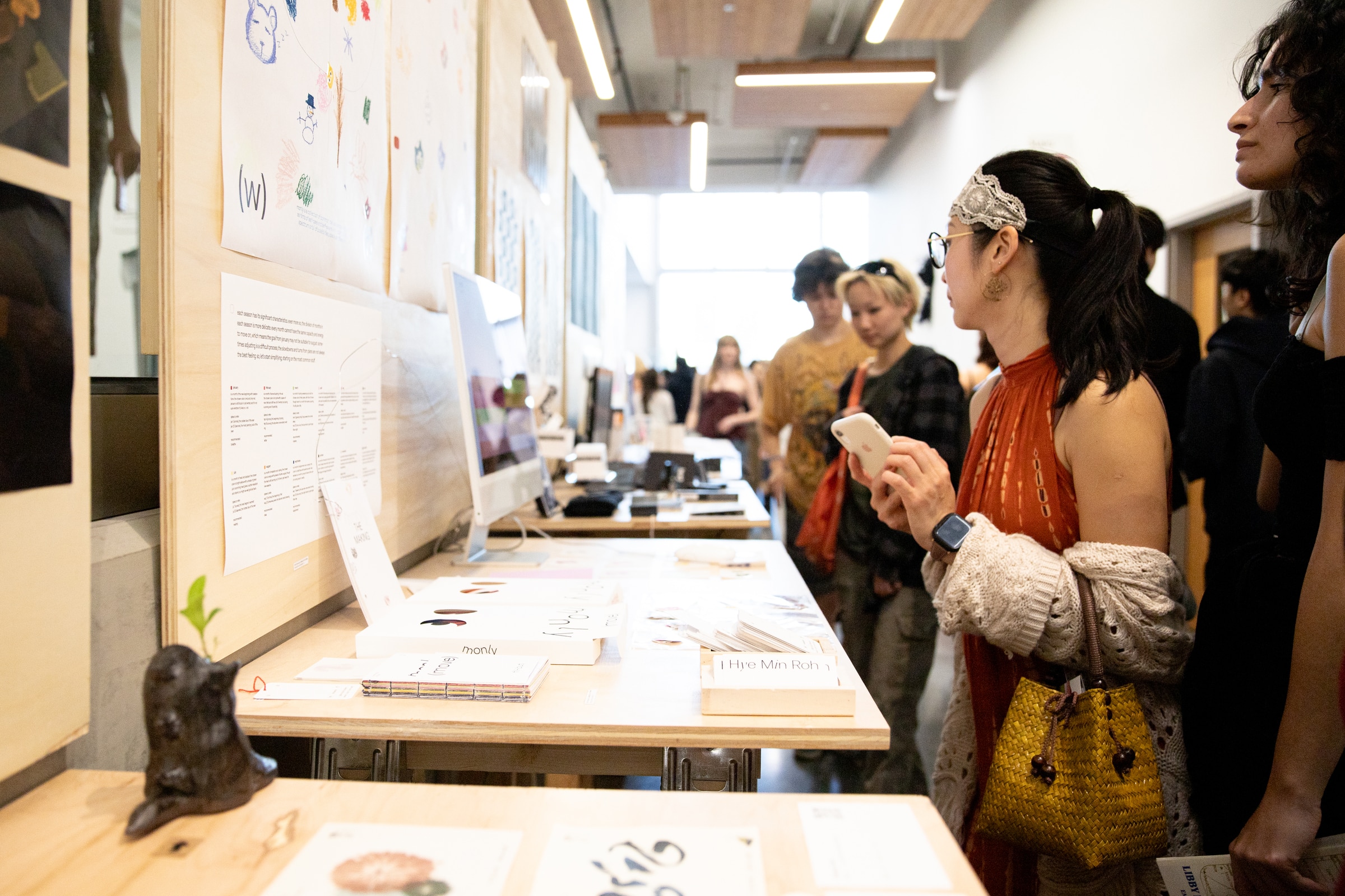 A woman holding a phone looks at student projects displayed on a long table and wall. Others stand nearby, examining prints, booklets, and digital work. The setting is a gallery-like exhibition space with natural light.
