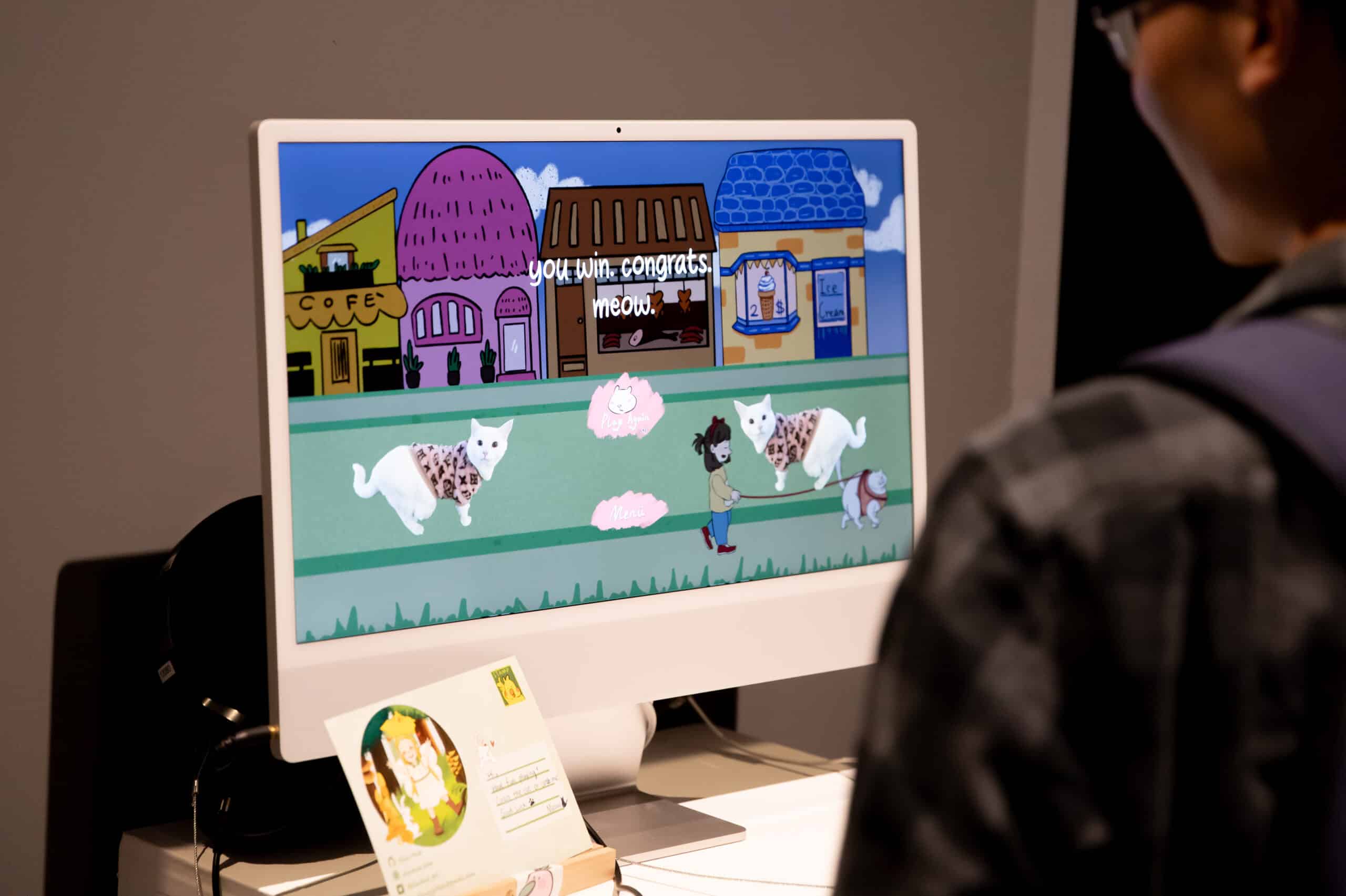 Person standing in front of a computer monitor displaying a colorful animated game scene with cats in sweaters and the text 'you win, congrats, meow.'