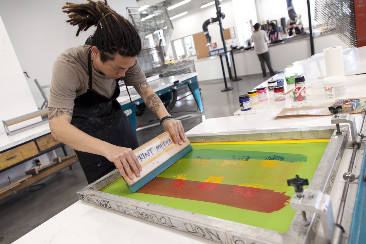 Person using a screen printing press in a studio, pulling ink across a screen with a squeegee labeled “PRINT MEDIA.” Bright colours and graphic shapes appear on the screen, while jars of ink and tools are spread across the worktable. Other people work in the background of the open studio space.