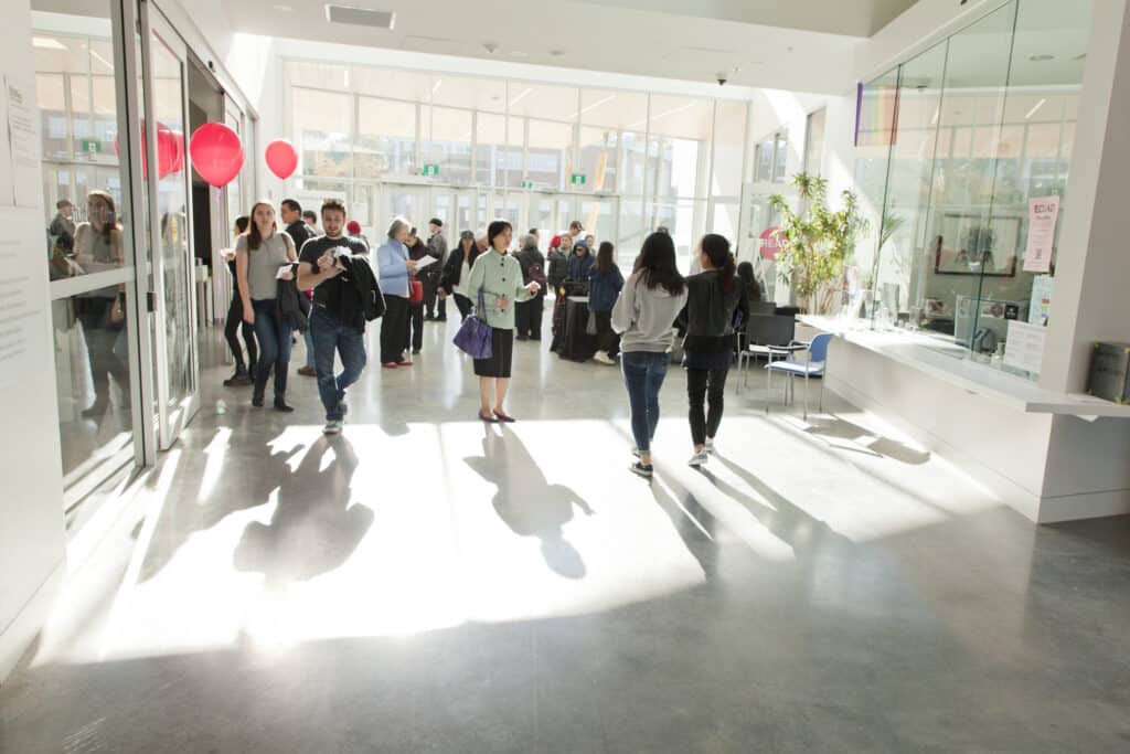 Bright, glass-walled campus lobby with sunlight streaming in as students and visitors mingle; a reception desk sits to the right and red balloons mark an event near the entrance.