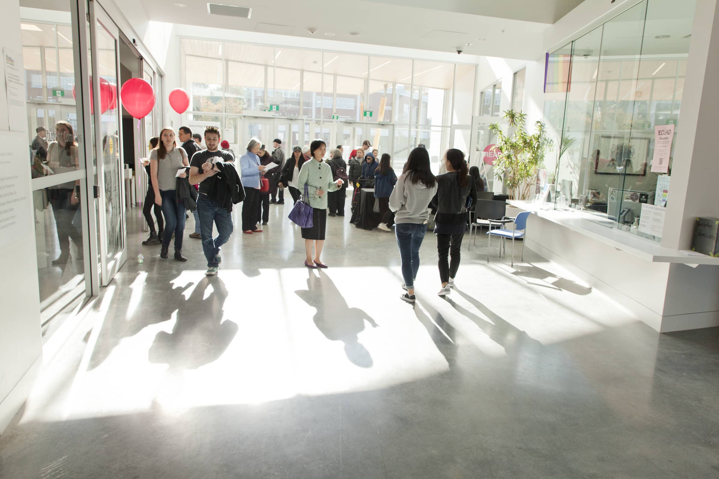 Bright, glass-walled campus lobby with sunlight streaming in as students and visitors mingle; a reception desk sits to the right and red balloons mark an event near the entrance.