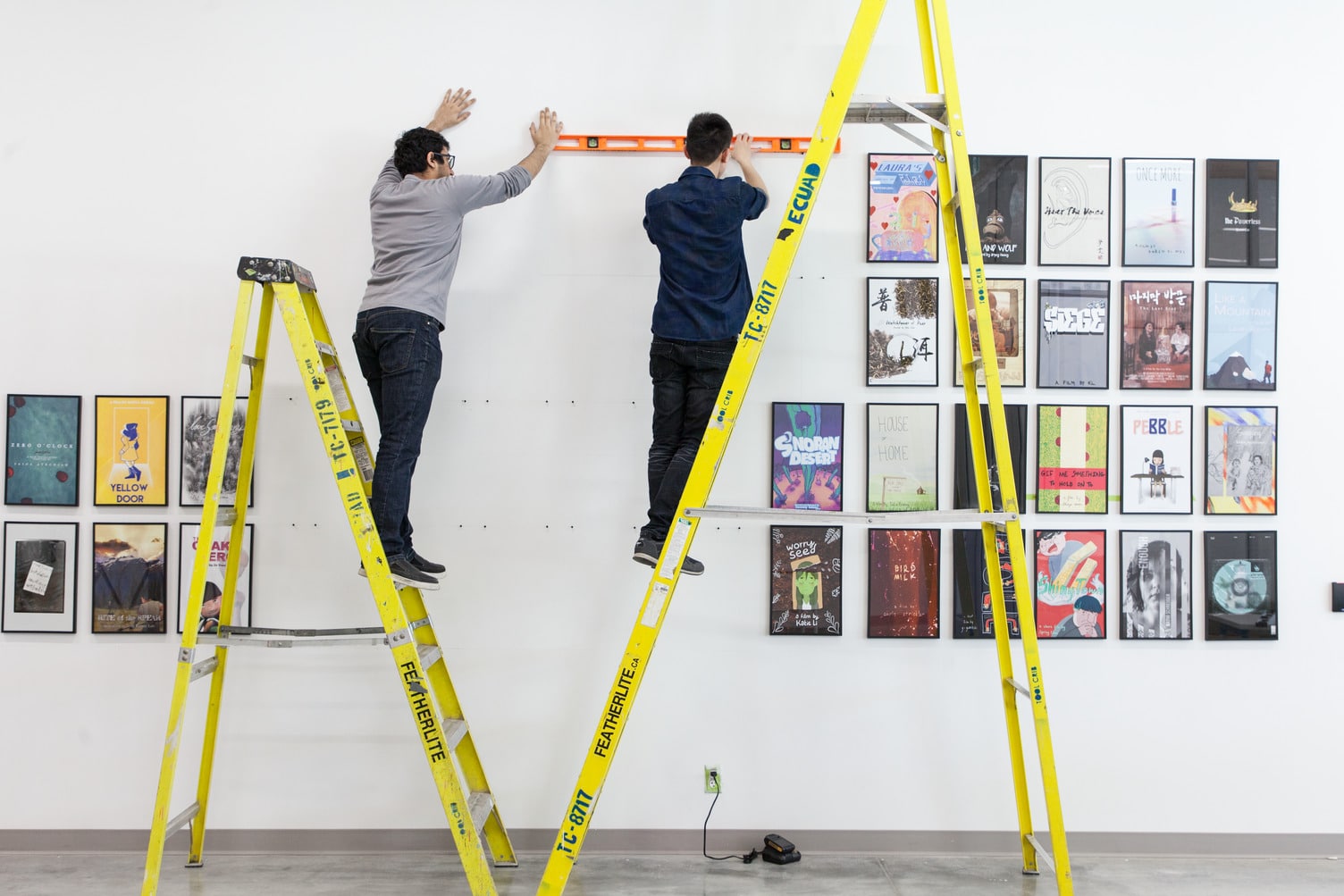 We see two tall, yellow ladders. There is a student on each one. Their backs are toward is as they use a level to hang a mosaic of framed posters on the wall.