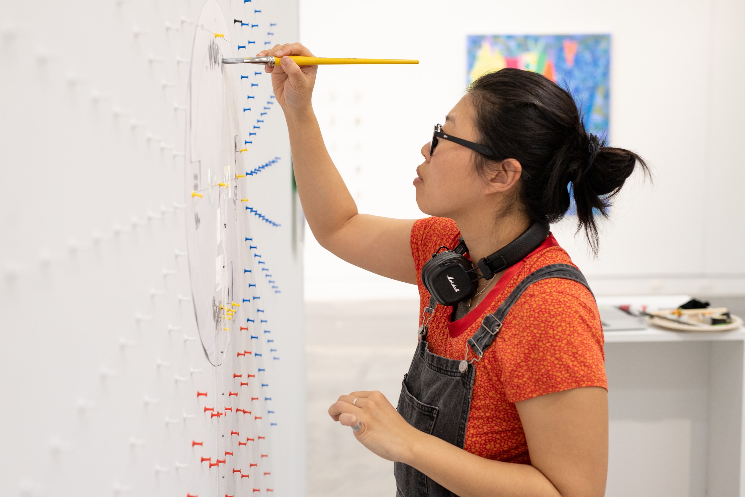Artist wearing glasses and headphones paints on a wall covered in colorful push pins arranged in patterns, part of an installation.