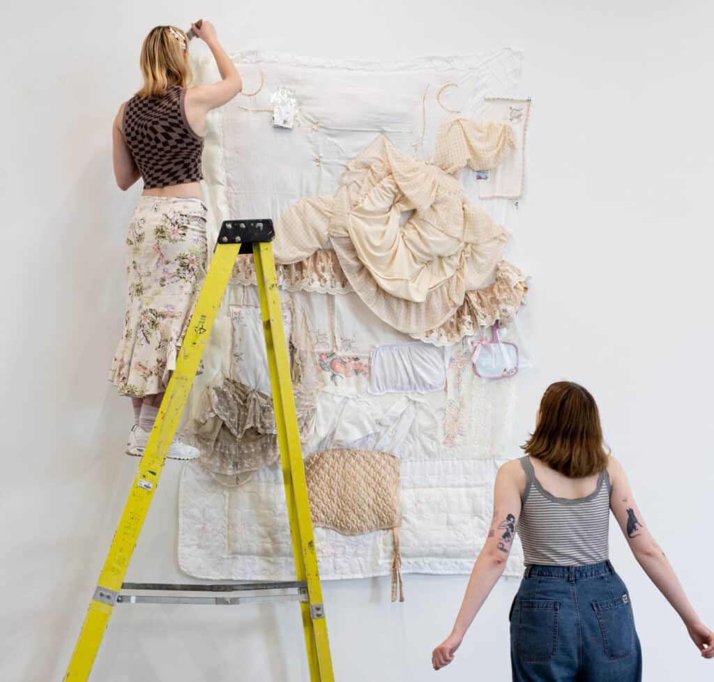 Two students install a large textile-based artwork on a gallery wall, one standing on a yellow ladder adjusting the piece while the other looks on from the floor.