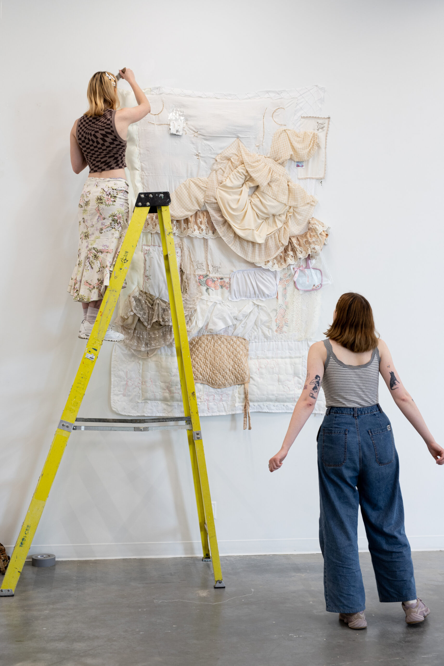 Two students install a large textile-based artwork on a gallery wall, one standing on a yellow ladder adjusting the piece while the other looks on from the floor.