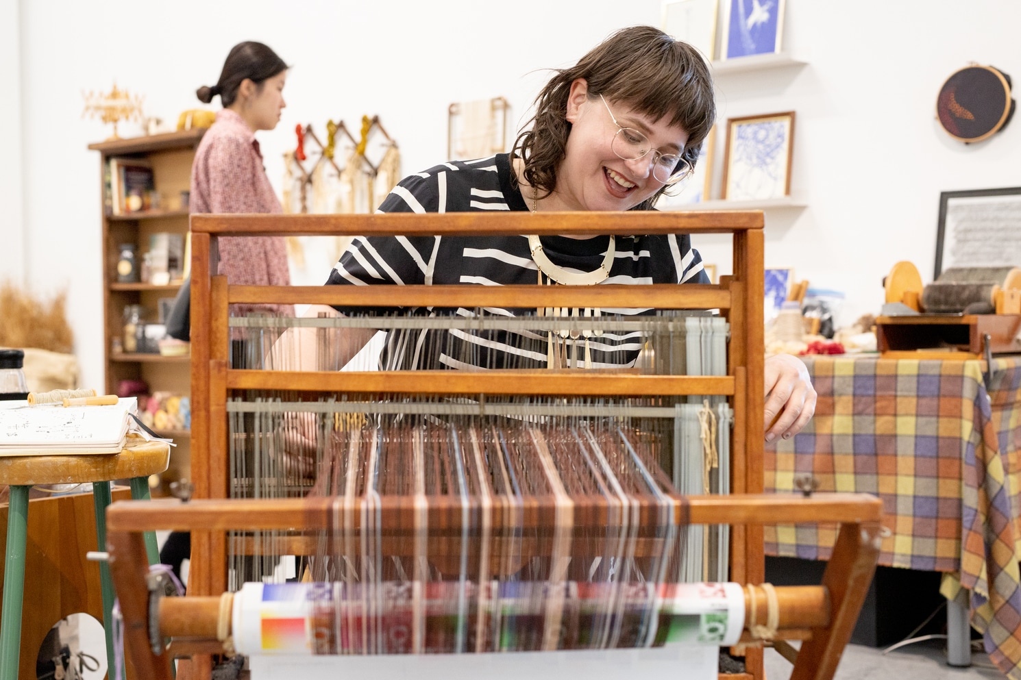 A smiling person wearing glasses and a striped shirt weaves at a large wooden loom, surrounded by craft tools and materials in a studio space.