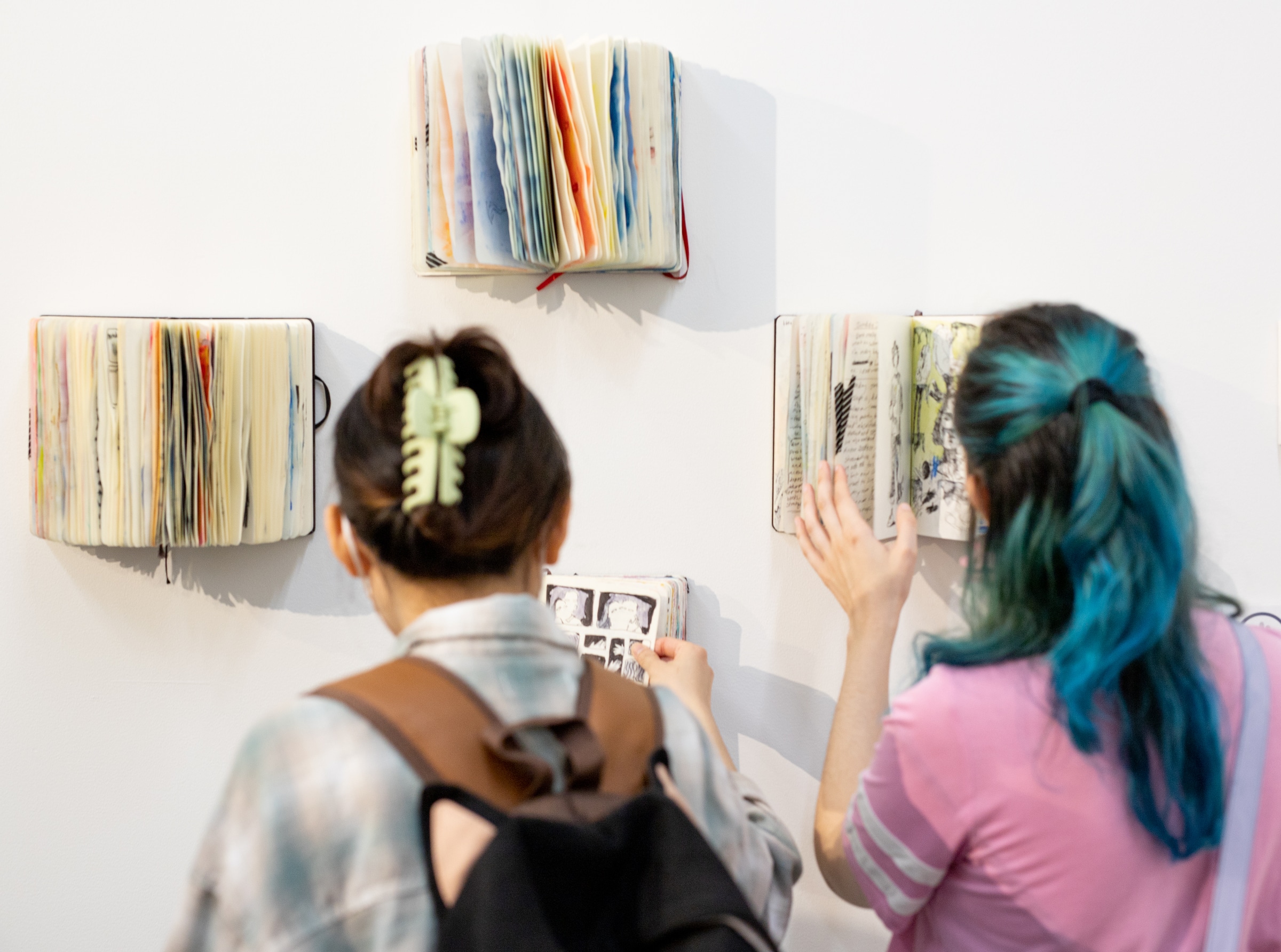 Two attendees at an art exhibition view books mounted on a white wall. From behind, one person with brown hair in a bun and a light green clip holds a small sketchbook with comic-style drawings. The other, with vibrant blue-green hair in a ponytail and a pink shirt, gently touches an open book displaying handwritten text and sketches. Several other colorful, fanned-out books are displayed on the wall as part of the installation.