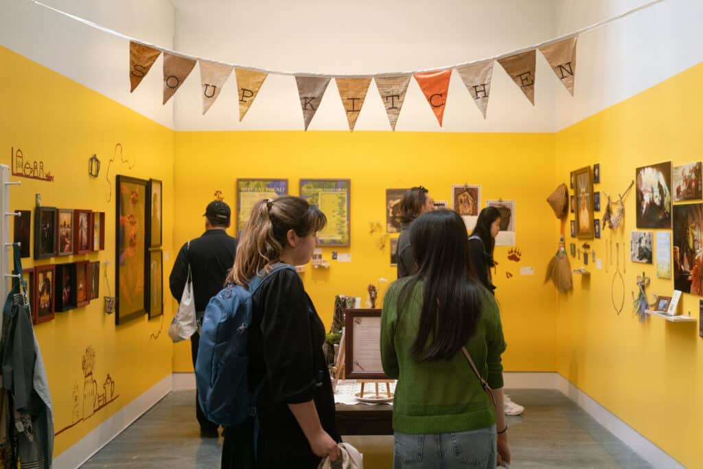 Using the movable walls, a small, enclosed section has been created in the Michael O'Brian Exhibition Commons. The small space has been painted bright yellow and the walls are covered with various artworks. Several people are in the space looking at them.