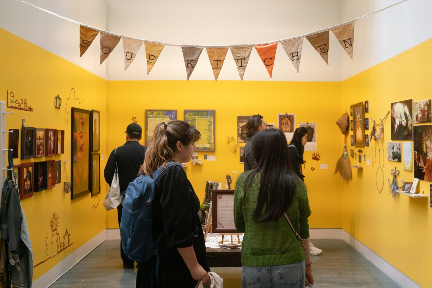 Using the movable walls, a small, enclosed section has been created in the Michael O'Brian Exhibition Commons. The small space has been painted bright yellow and the walls are covered with various artworks. Several people are in the space looking at them.