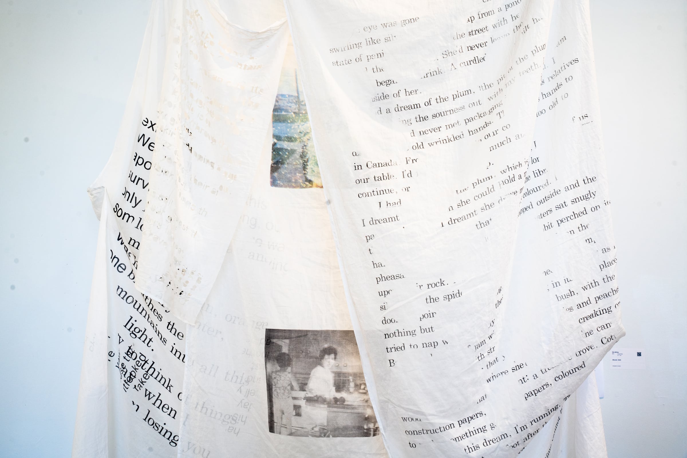 White fabric panels hang loosely, printed with large blocks of black text and faded photographs. The installation creates a layered, draped environment with overlapping text and imagery.
