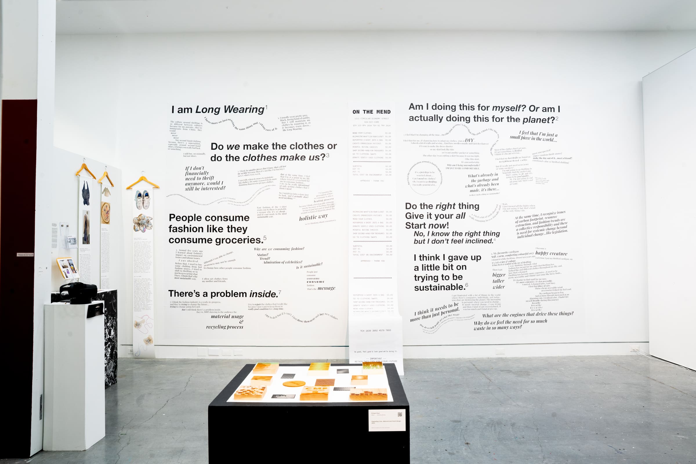 Art installation in a gallery featuring two large white wall panels covered in bold black text and handwritten notes about sustainability, clothing, and consumer habits. Smaller artworks with hanging garments are displayed on the left, with a black pedestal in the foreground showing samples and text.