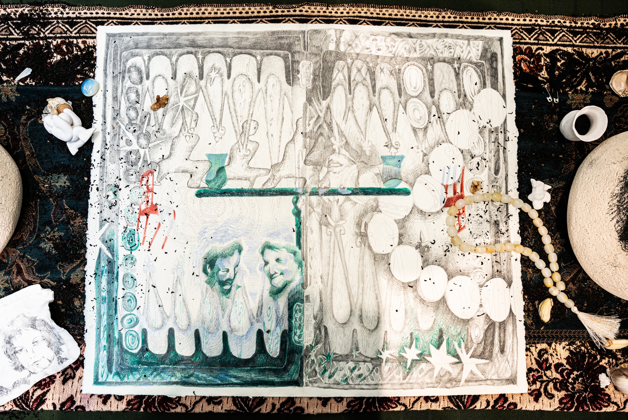 Mixed-media artwork on paper displayed with small objects around it, featuring pencil drawings, green accents, portraits, and layered textures.