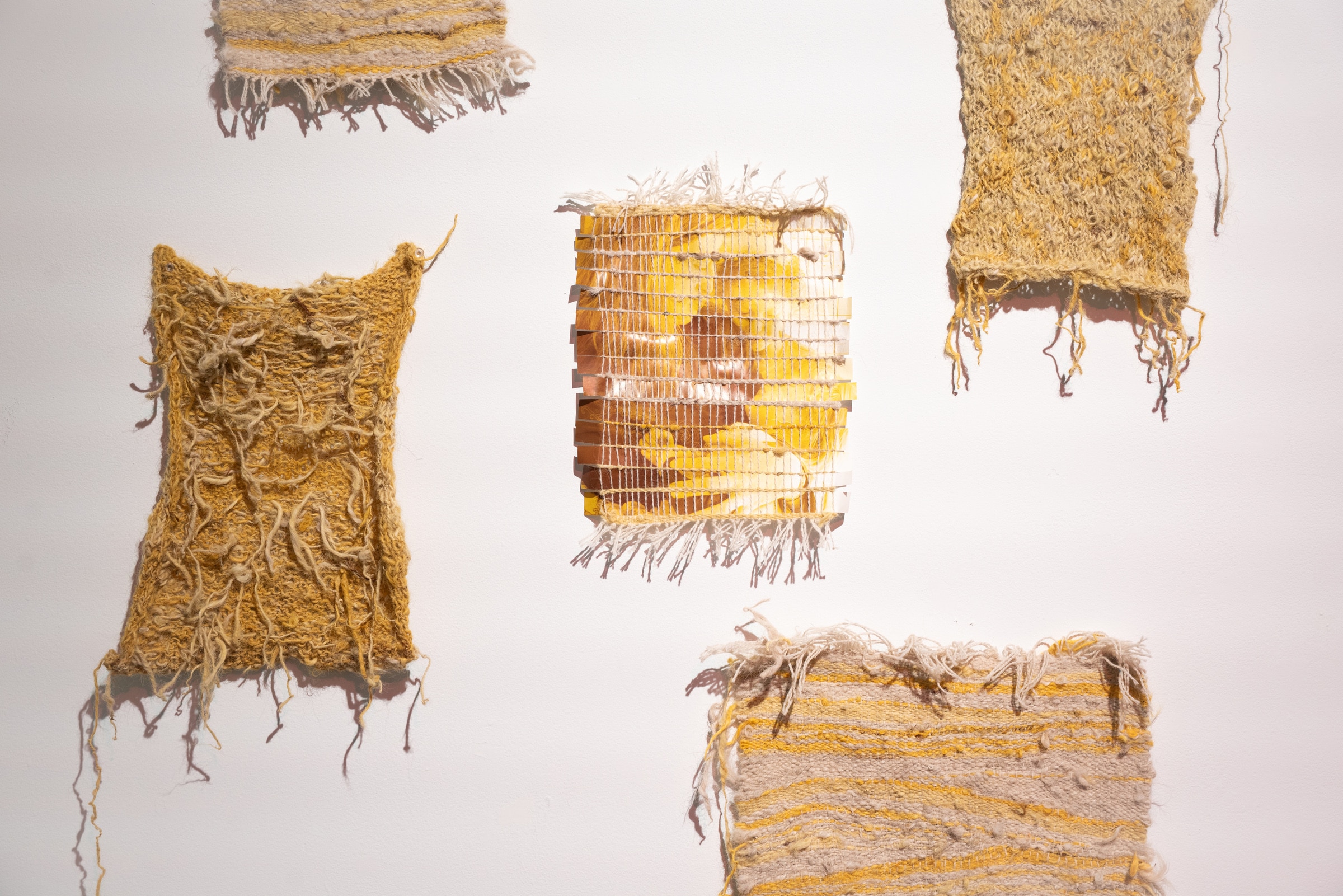 Handwoven textile pieces in shades of yellow, beige, and cream displayed on a white wall, featuring varied textures from smooth woven patterns to rough, knotted fibers.