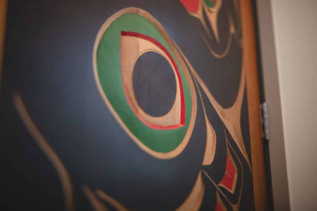 Close-up of an Indigenous Northwest Coast-style artwork featuring a carved circular form with green, red, and natural wood tones on a dark background.