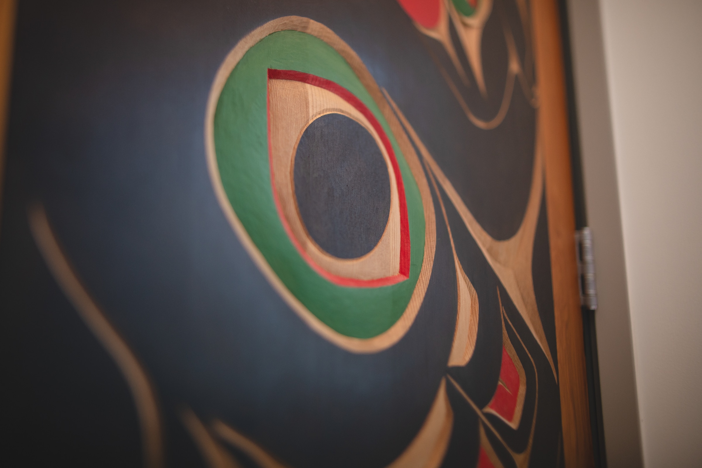 Close-up of an Indigenous Northwest Coast-style artwork featuring a carved circular form with green, red, and natural wood tones on a dark background.