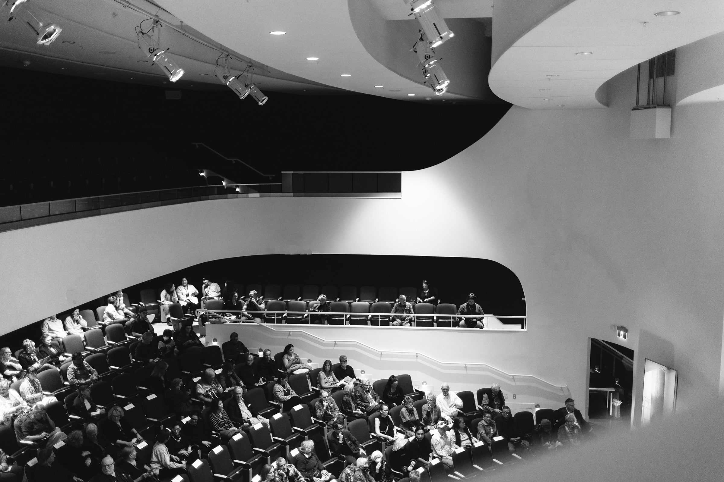 Audience seated in a modern, multi-tiered auditorium with curved walls and upholstered chairs, waiting for an event to begin under ceiling spotlights.