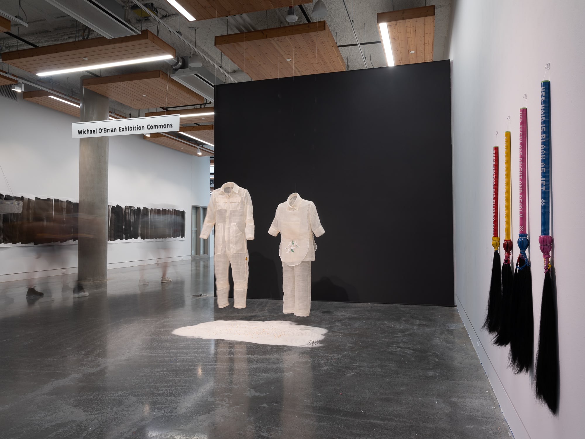 Two translucent garment sculptures, one adult-sized and one child-sized, hang above a bed of sand, with four braided hair pieces mounted on the wall.