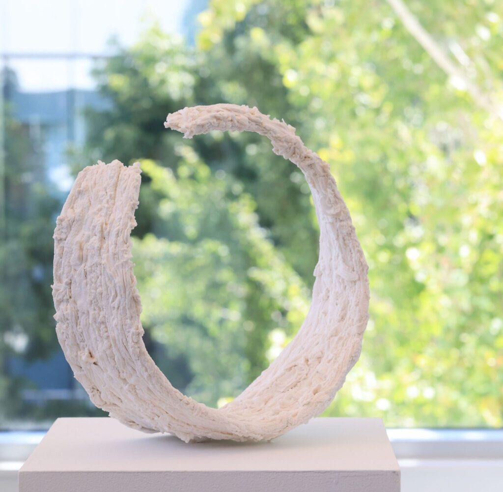 A white, textured circular sculpture resembling an open ring, made from layered paper clay, displayed on a pedestal in front of a window with trees visible outside.