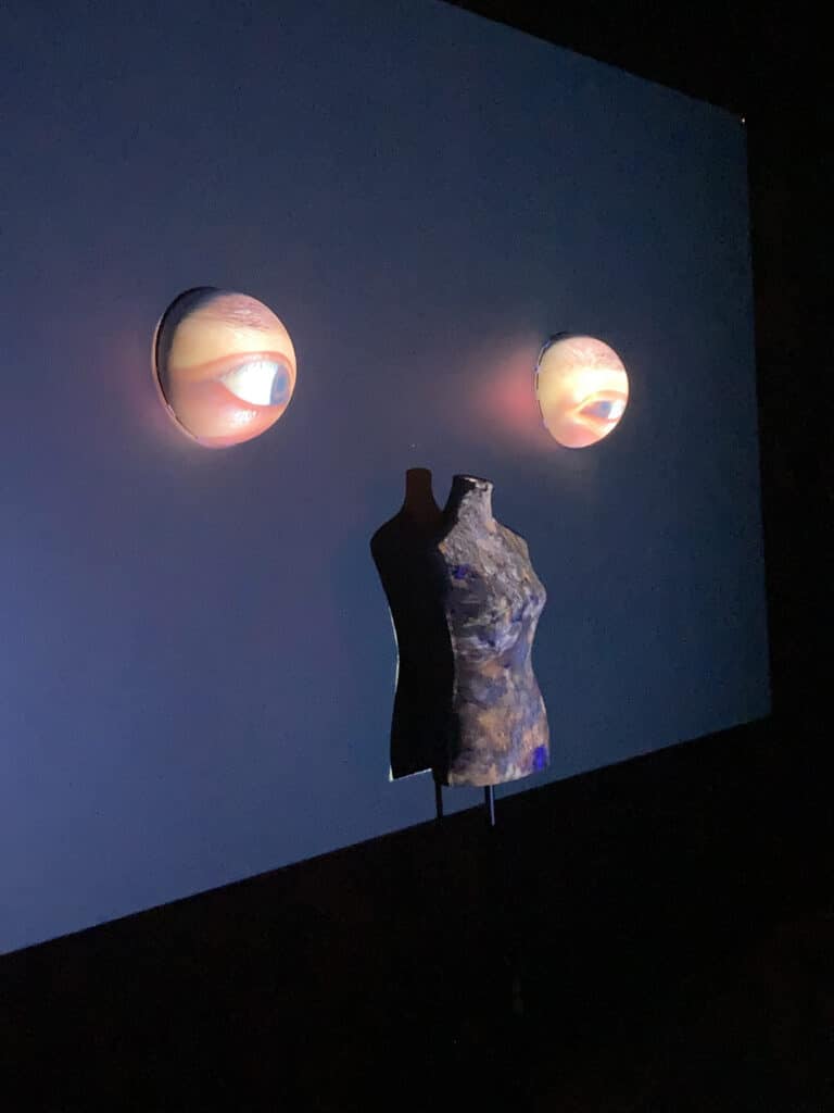 An art installation featuring a partial mannequin torso mounted on a stand, with two glowing projections of human eyes on a dark wall behind it.