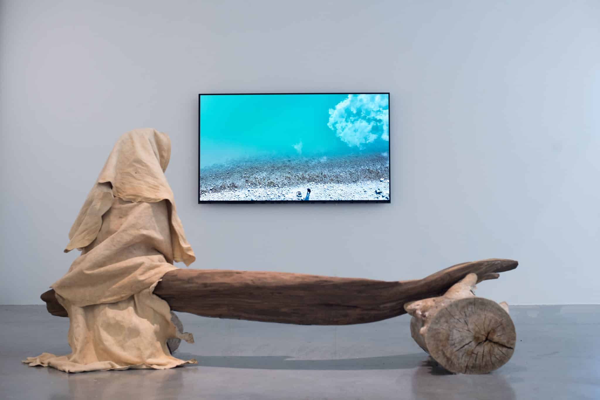 A draped fabric figure sits on a large log sculpture in front of a video screen showing an underwater seascape with turquoise water and sand.