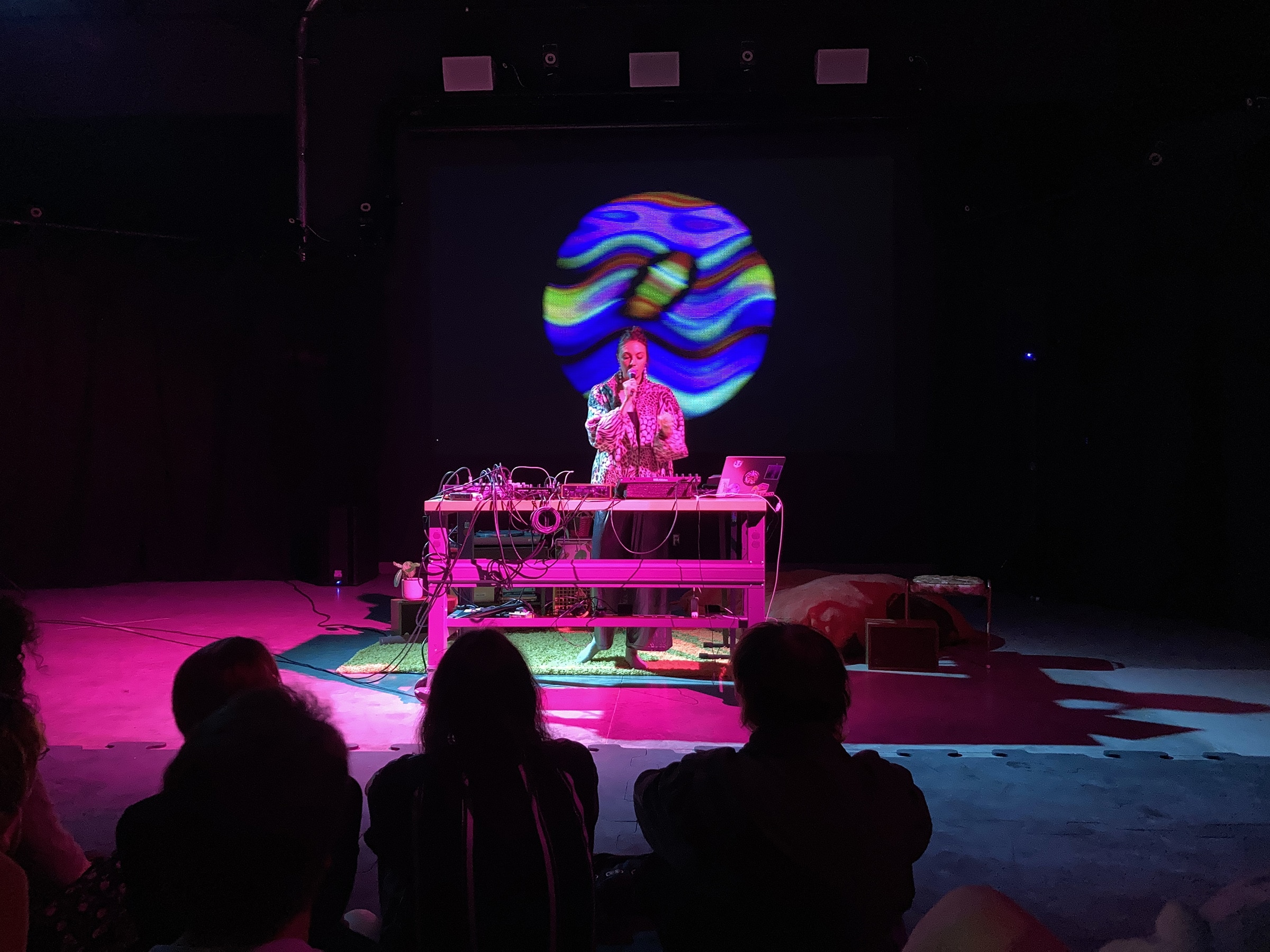 A performer stands behind a table of electronic music equipment, speaking into a microphone, with a swirling, colorful globe-like projection displayed behind them; audience members sit on the floor in front.