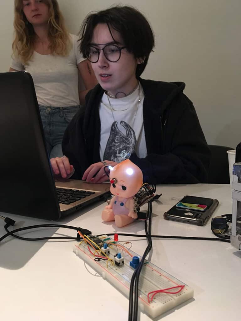 A student works on a laptop while an electronic toy baby doll sits in front of them, glowing with LED lights wired to a breadboard circuit. Another student stands in the background watching.