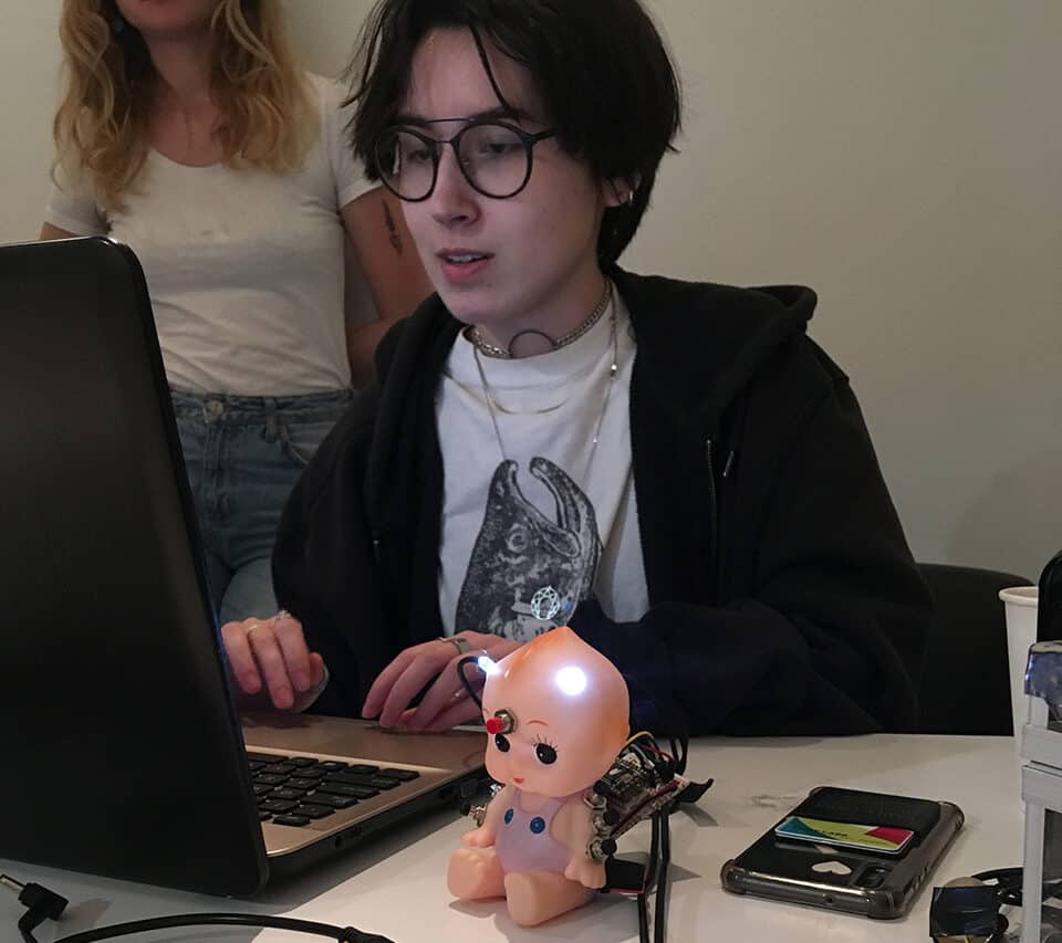 A student works on a laptop while an electronic toy baby doll sits in front of them, glowing with LED lights wired to a breadboard circuit. Another student stands in the background watching.