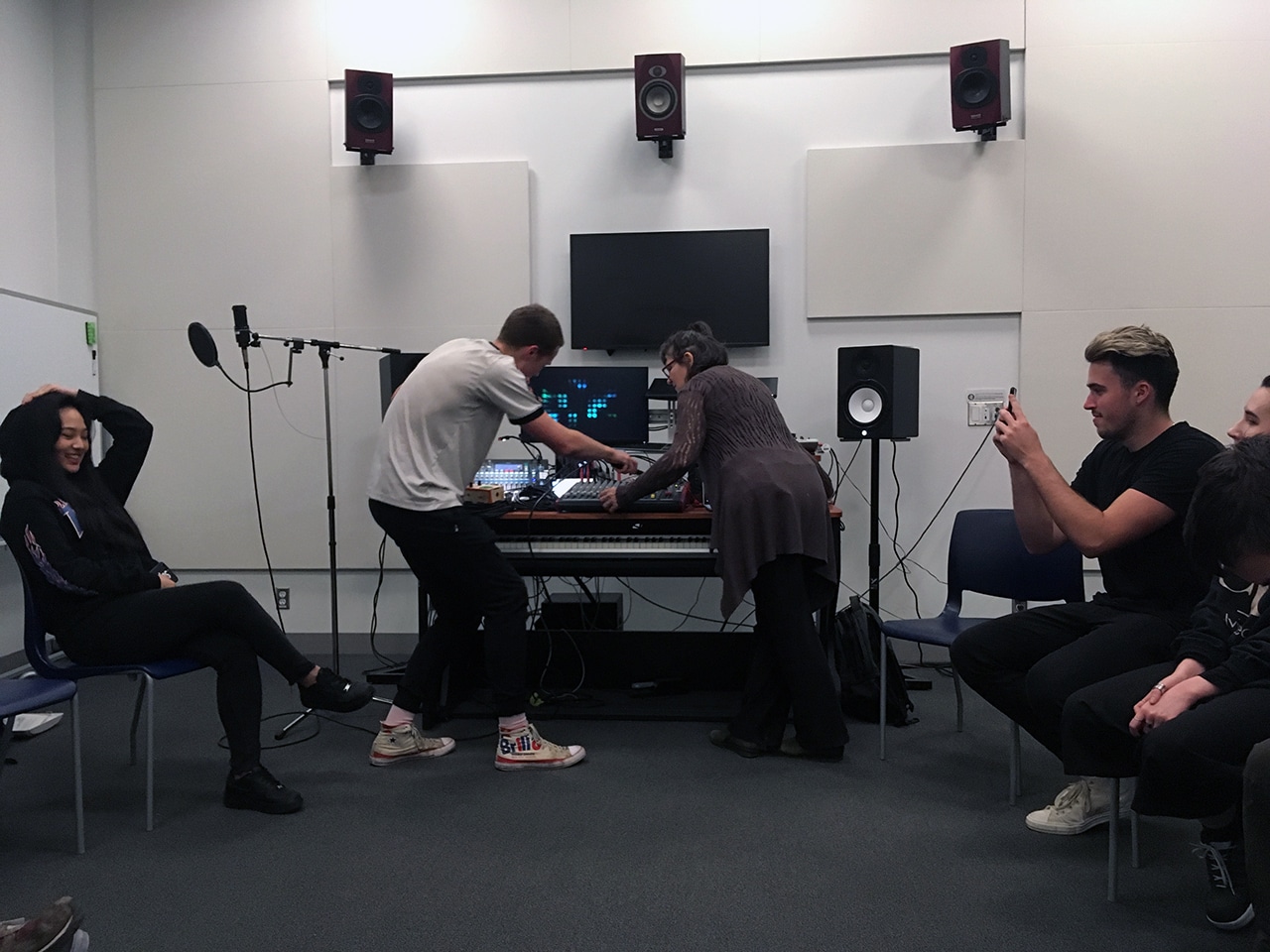 Students gather in a sound lab, with one adjusting audio equipment while another records with a phone; microphones, speakers, and a mixing console are set up in the room.