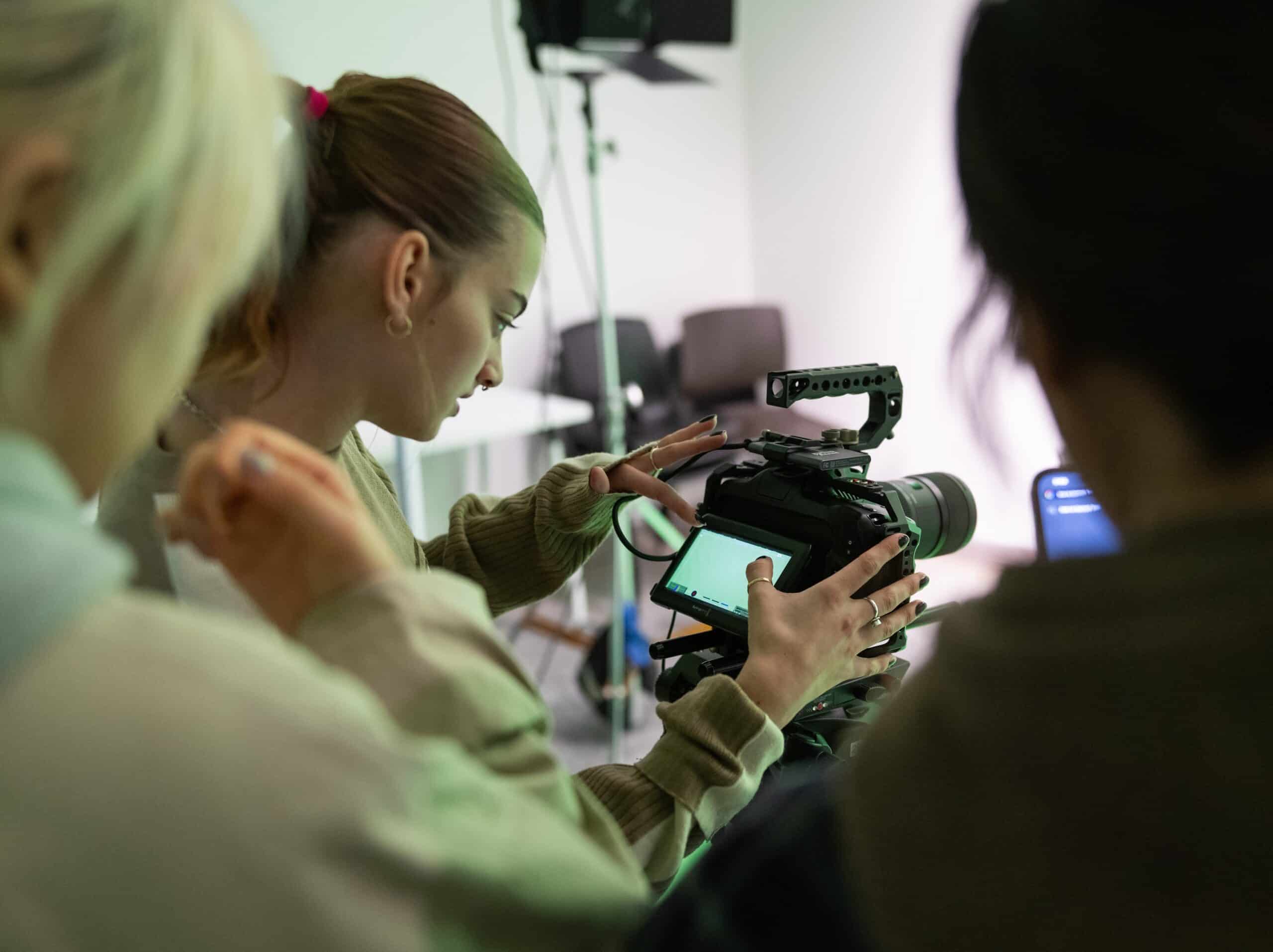 A group of students gathers around a professional video camera in a studio setting. One student adjusts the camera’s touchscreen while others look on, learning how to operate the equipment.