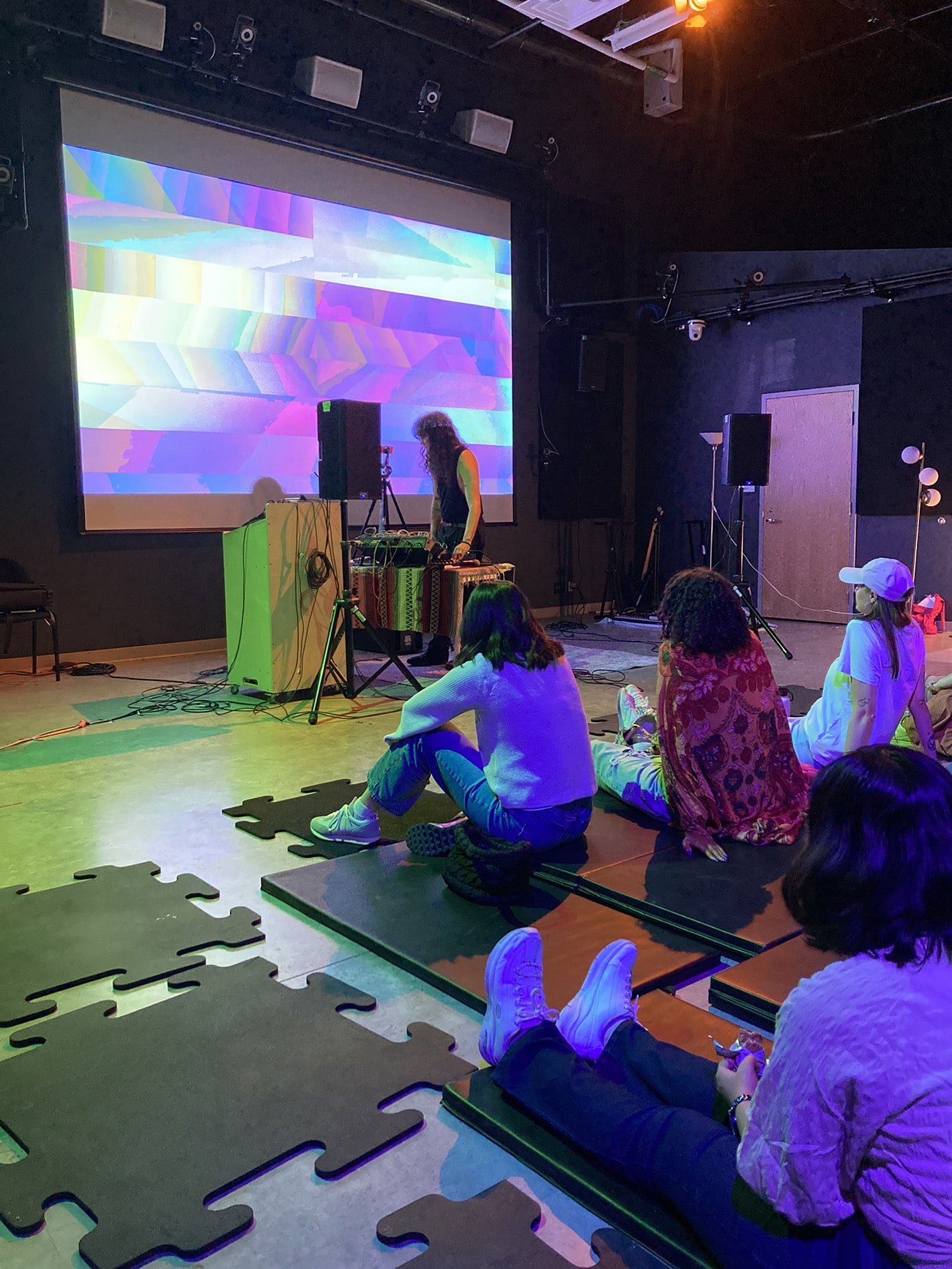 A performer operates sound equipment in front of a large projection of colorful, glitch-like digital graphics, while several students sit on mats on the floor watching.