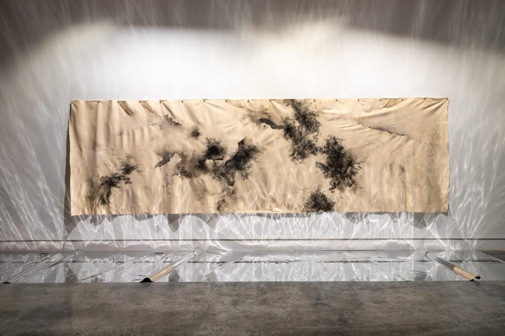 Large tan canvas with black ink-like marks hanging on a gallery wall, with reflective silver material on the floor casting shimmering light patterns across the installation.