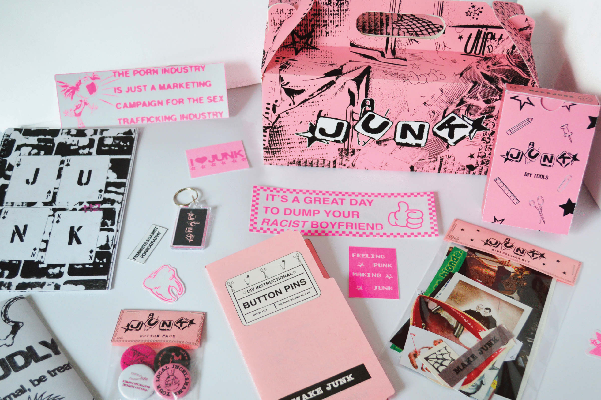A collection of pink and black DIY punk-inspired merchandise arranged on a white surface. Items include a pink cardboard carrying case with the word “JUNK” spelled out in cutout letters, booklets, keychains, stickers, buttons, and zines. Some text on the items reads: “The porn industry is just a marketing campaign for the sex trafficking industry,” “It’s a great day to dump your racist boyfriend,” “Feeling punk making junk,” and “Button Pins.” The overall aesthetic is bold, activist, and DIY.