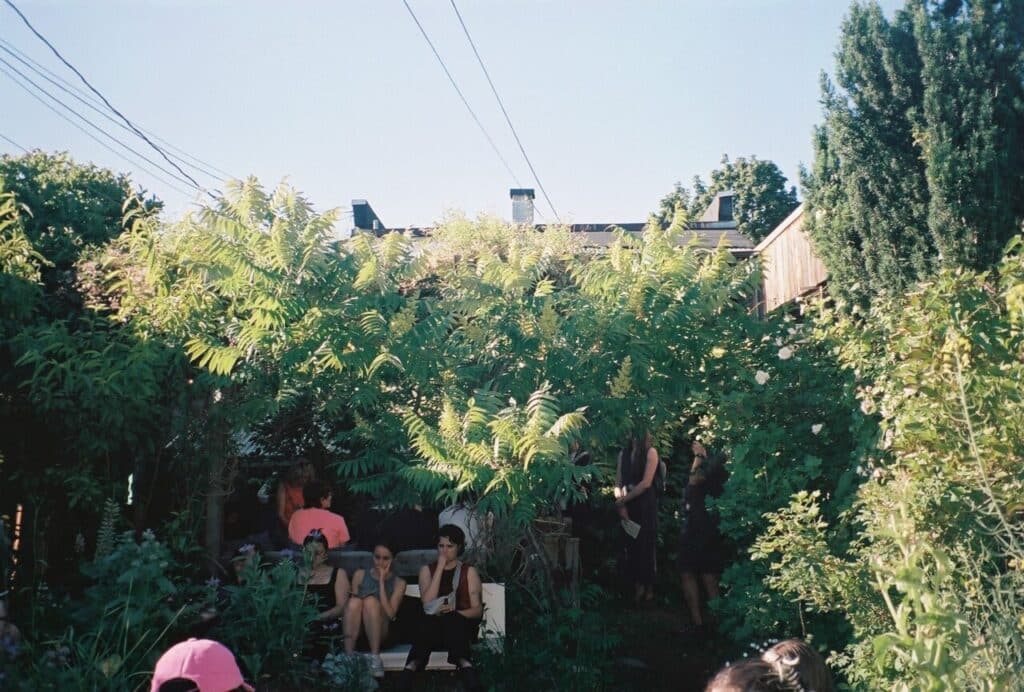 A group of people sit and stand under dense green foliage in a backyard, partially hidden by leaves and branches.