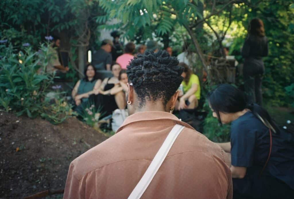 The back of a person wearing a brown shirt, looking toward a group of seated people in a shaded garden.