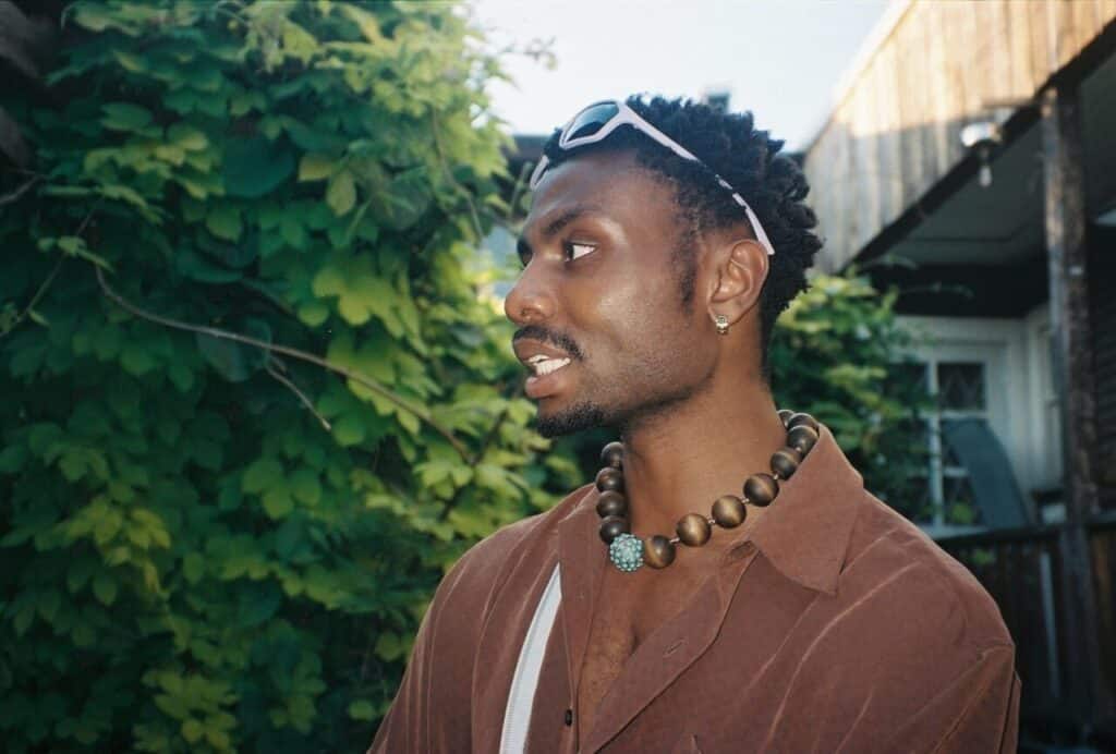 A close-up side profile of a person in a brown shirt, sunglasses on their head, and a beaded necklace, standing near lush greenery.
