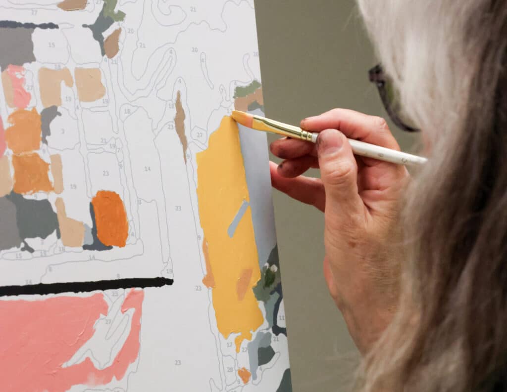 Close-up of a hand painting a numbered canvas with a small brush, filling a section with bright yellow paint.